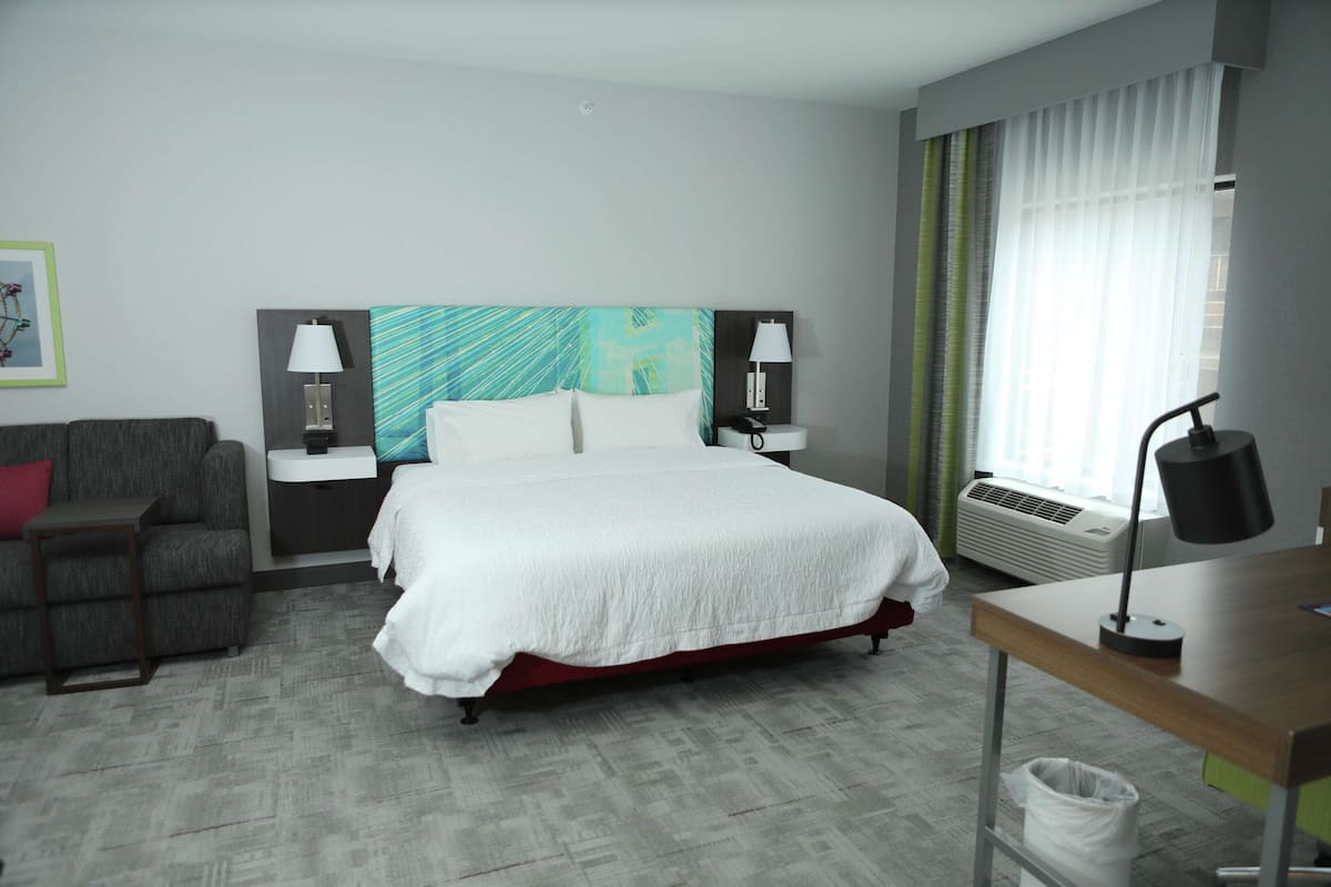 Une chambre d’hôtel comprenant un lit, un bureau, une chaise, une table de chevet, une lampe, un cadre photo et une fenêtre avec des rideaux.