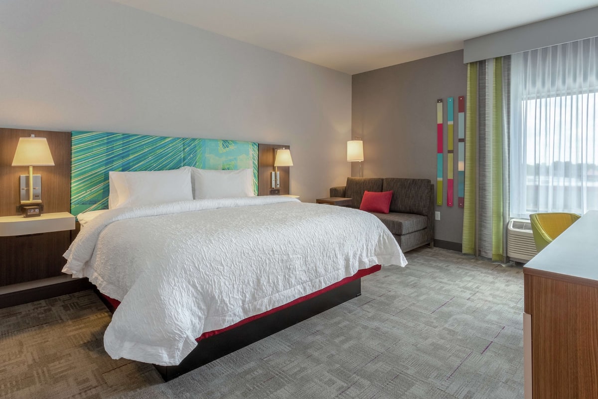 Une chambre d’hôtel avec un grand lit, deux tables de chevet avec des lampes, un canapé avec des coussins, un tableau abstrait aux couleurs vives accroché au mur, et une fenêtre avec des rideaux légers.