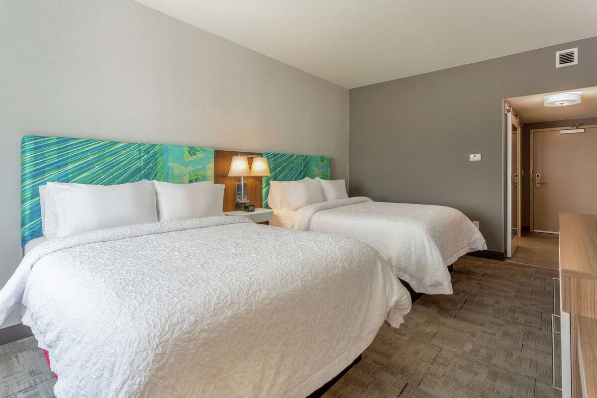 Une chambre d’hôtel avec deux lits, une tête de lit ornée d’un motif tropical, une table de chevet avec une lampe et une porte donnant sur une autre pièce.