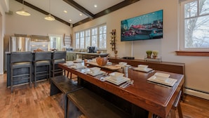 Una cocina moderna con comedor, vigas de madera, ventanas amplias y un televisor que muestra un paisaje urbano.