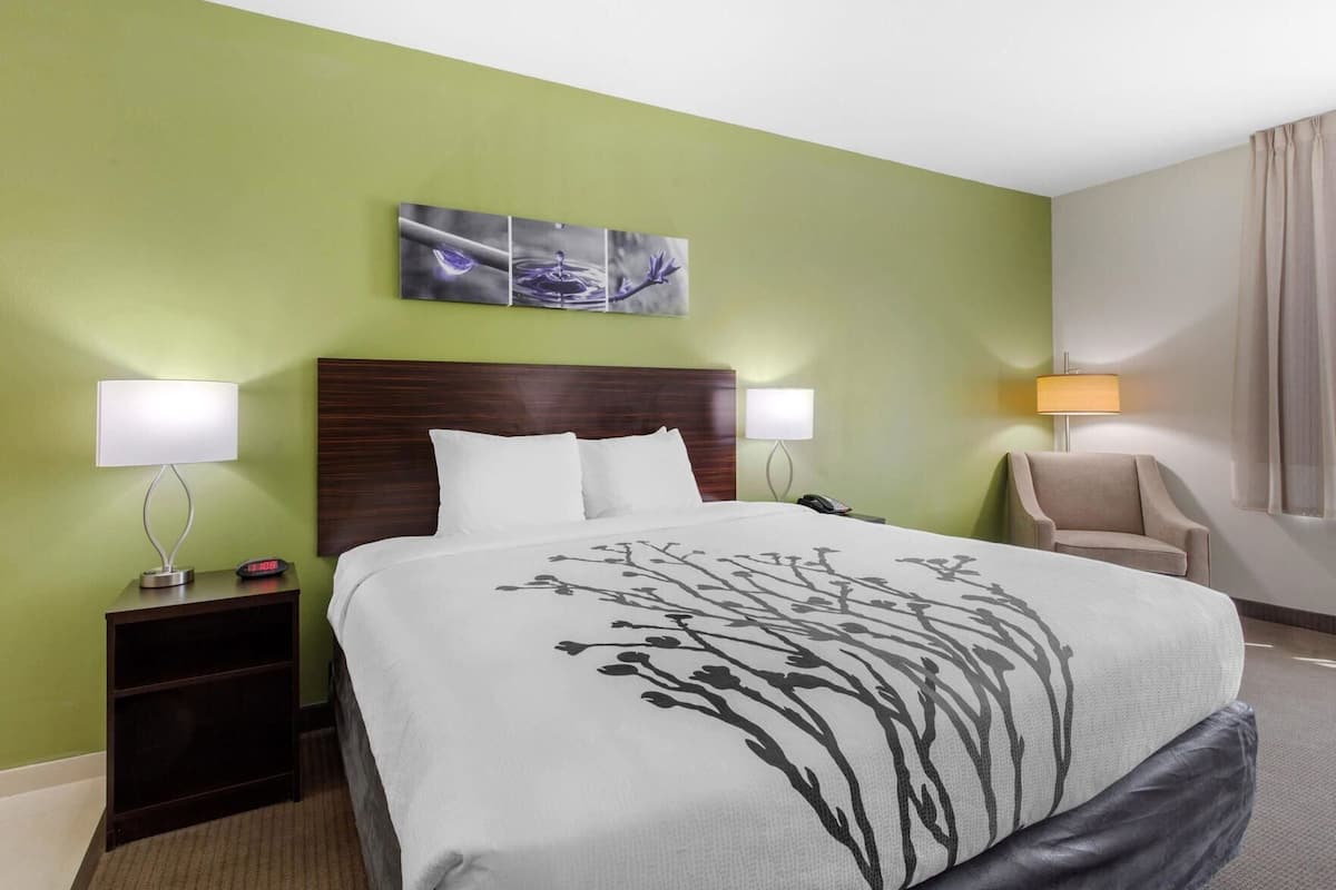 Ein Hotelzimmer mit einem Bett, zwei Nachttischlampen, einem Nachttisch, einem Sessel, einem Bild an der Wand und einem Fenster mit Vorhängen.