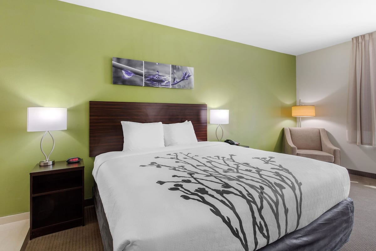 Ein Hotelzimmer mit einem großen Bett, zwei Nachttischen mit Lampen, einem Sessel und einem an der Wand hängenden Kunstwerk.