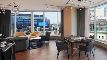 Een moderne hotelkamer met een groot raam met uitzicht op de stad, een gezellige zithoek met een bank en fauteuils, een eettafel met een schaakbord en een klein barretje.