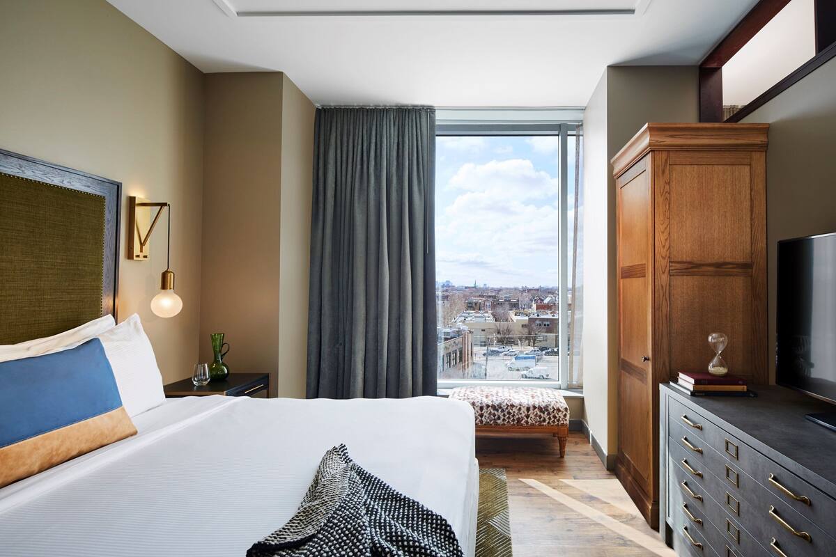 Habitación de hotel con una cama grande, un armario de madera, un televisor y vista a la ciudad a través de una ventana.