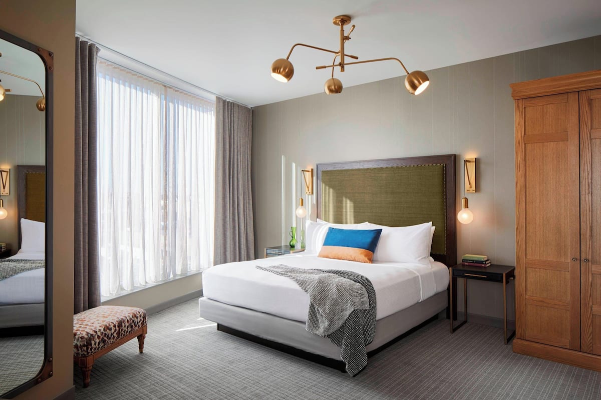 Habitación de hotel con una cama grande, un armario de madera, una mesita de noche con lámpara y un ventanal con cortinas.