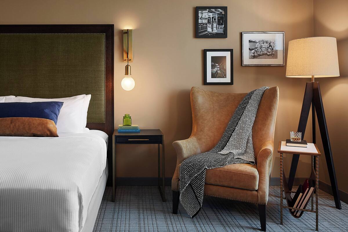 Una habitación de hotel con una cama, una silla, una mesita de noche, una lámpara y cuadros en la pared.
