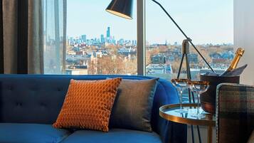 Uma sala de estar moderna com um sofá azul, uma janela grande com vista para a cidade, um abajur preto e uma mesa redonda pequena com uma garrafa e copos.
