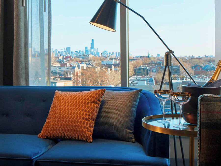 Uma sala de estar moderna com um sofá azul, uma janela grande com vista para a cidade, um abajur preto e uma mesa redonda pequena com uma garrafa e copos.