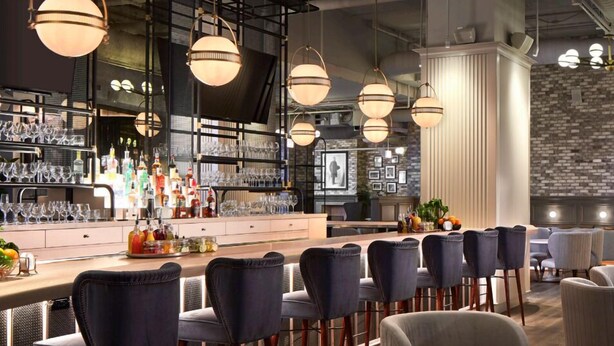 A bar area with multiple seating options, including bar stools and round tables, with a variety of bottles and glasses on the bar counter. - Hotel Zachary Chicago, a Tribute Portfolio Hotel (Chicago)