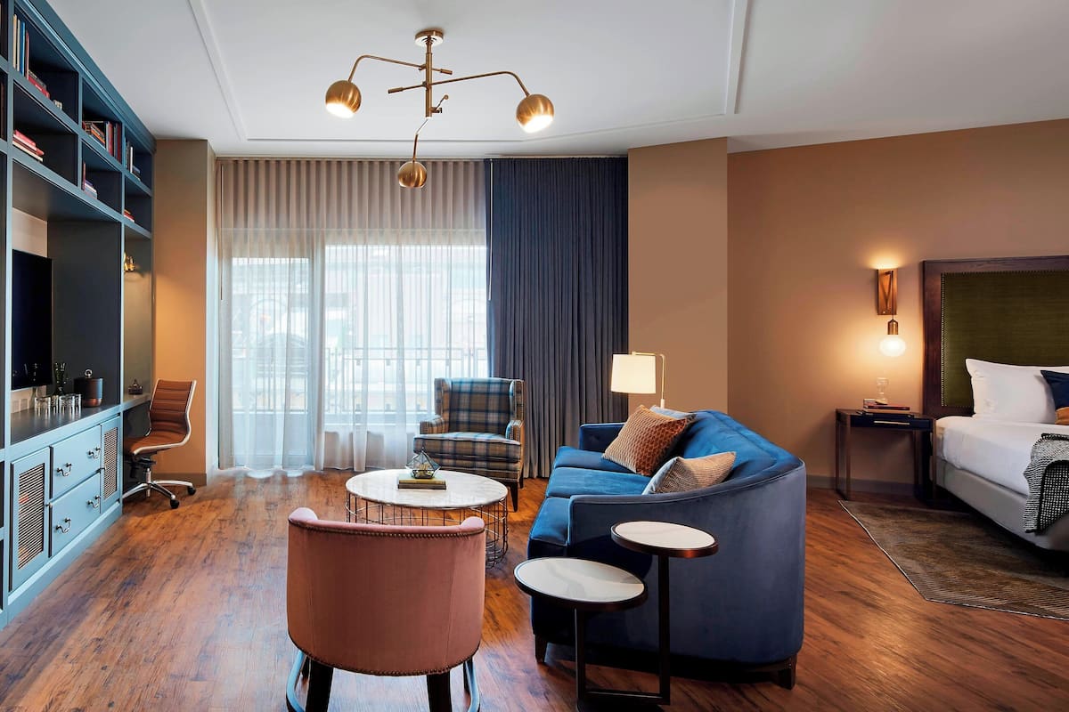 Una habitación de hotel con un sofá azul, una mesa de centro redonda, un sillón rosa, una estantería, un escritorio y una cama con cabecera.