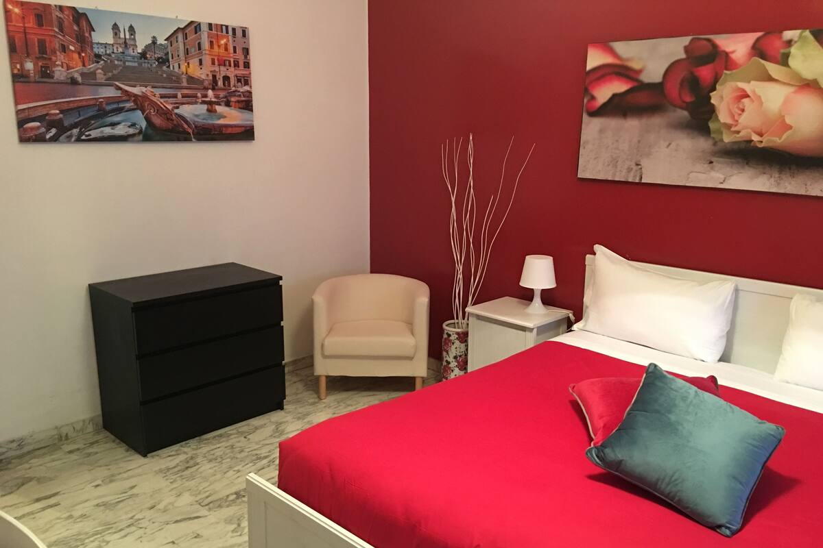 A bedroom with a red bedspread, a white headboard, a black dresser, a beige armchair, a nightstand with a lamp, and two paintings on the walls.