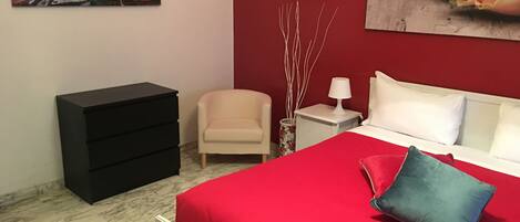 A bedroom with a red bedspread, a white headboard, a black dresser, a beige armchair, a nightstand with a lamp, and two paintings on the walls.