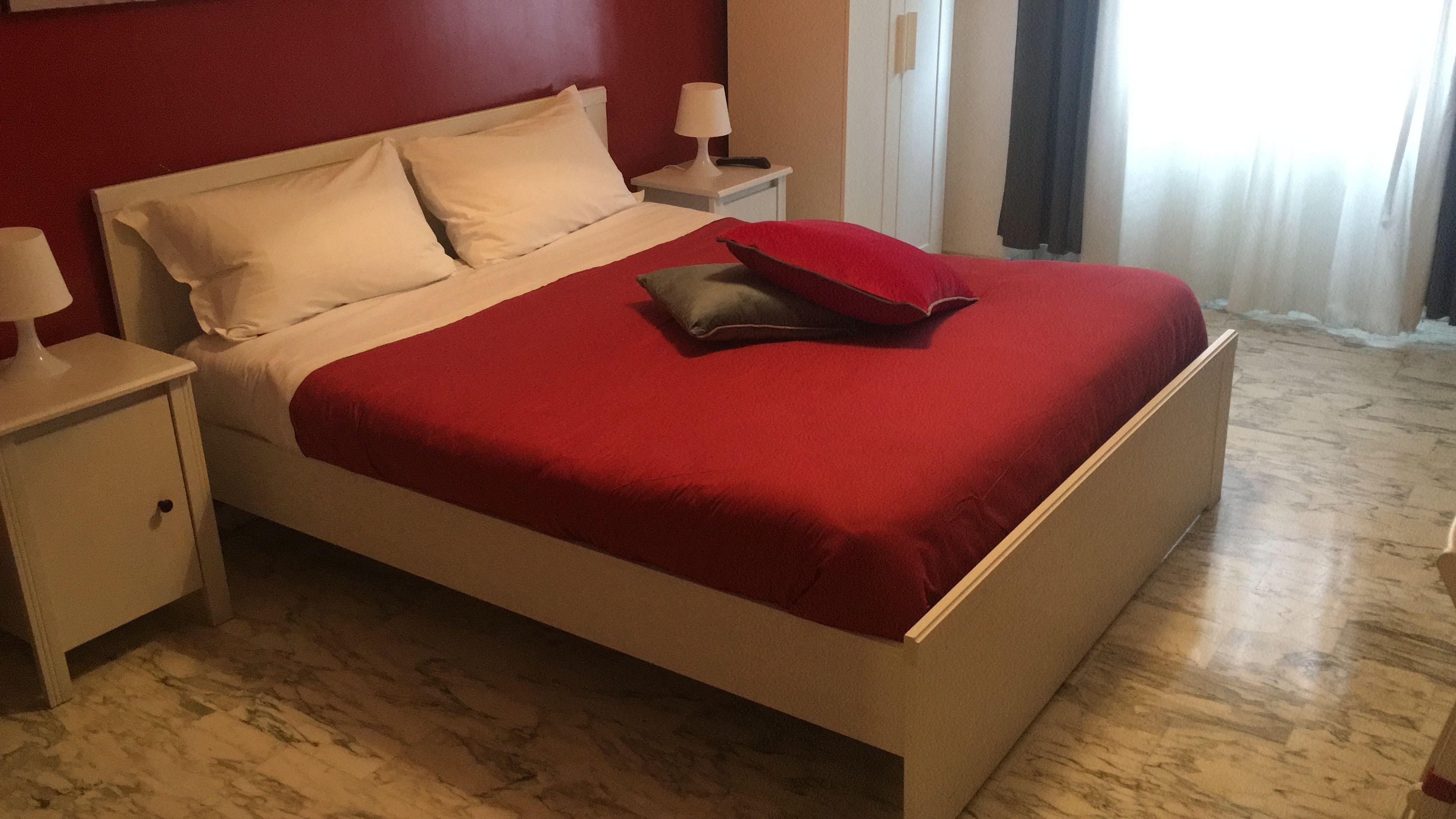 Una cama bien hecha con ropa de cama blanca y roja, un cojín decorativo rojo y una mesita de noche con una lámpara.