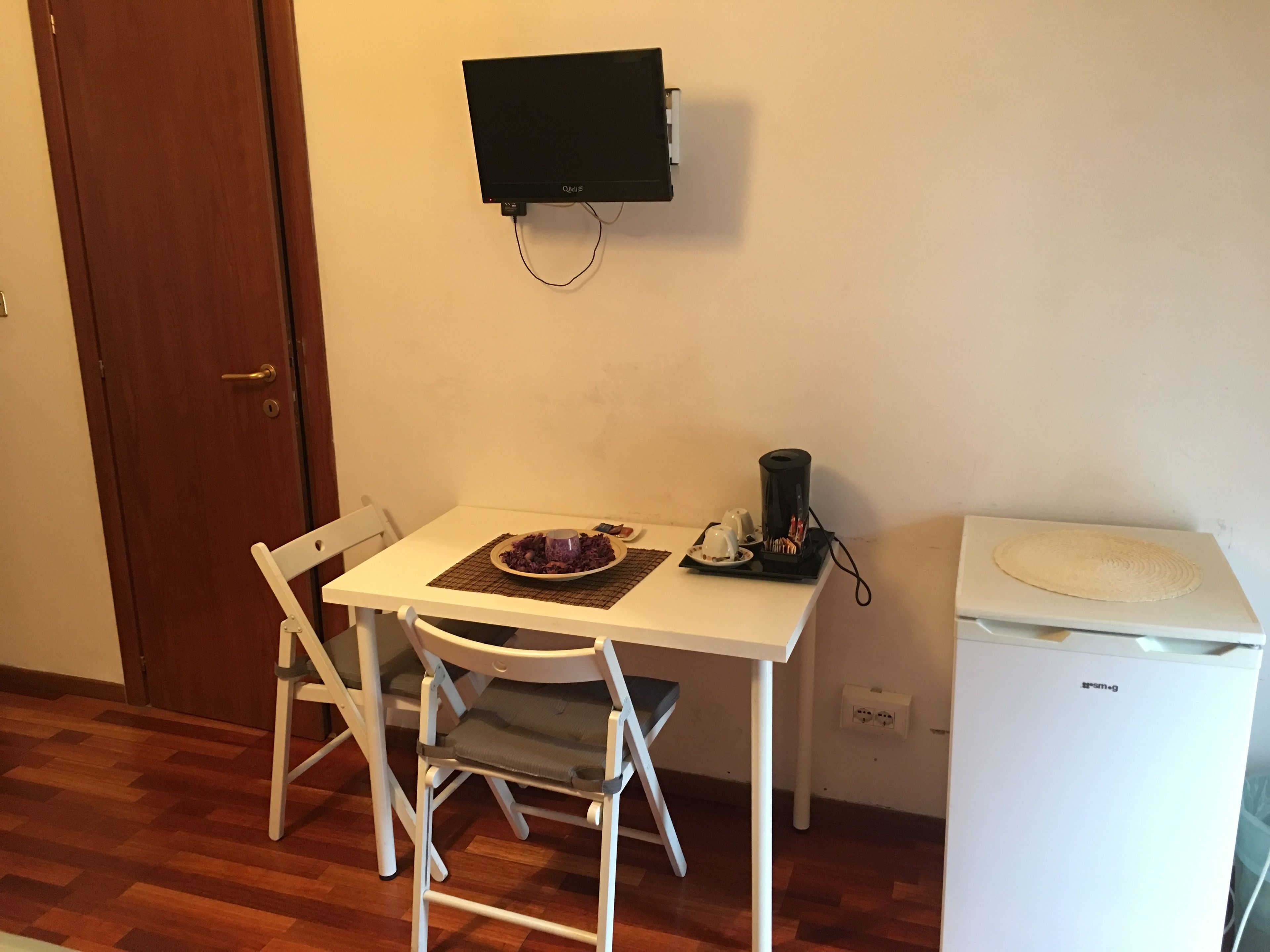 Une petite pièce avec une table blanche, deux chaises, un téléviseur noir fixé au mur, une machine à café et une poubelle blanche.