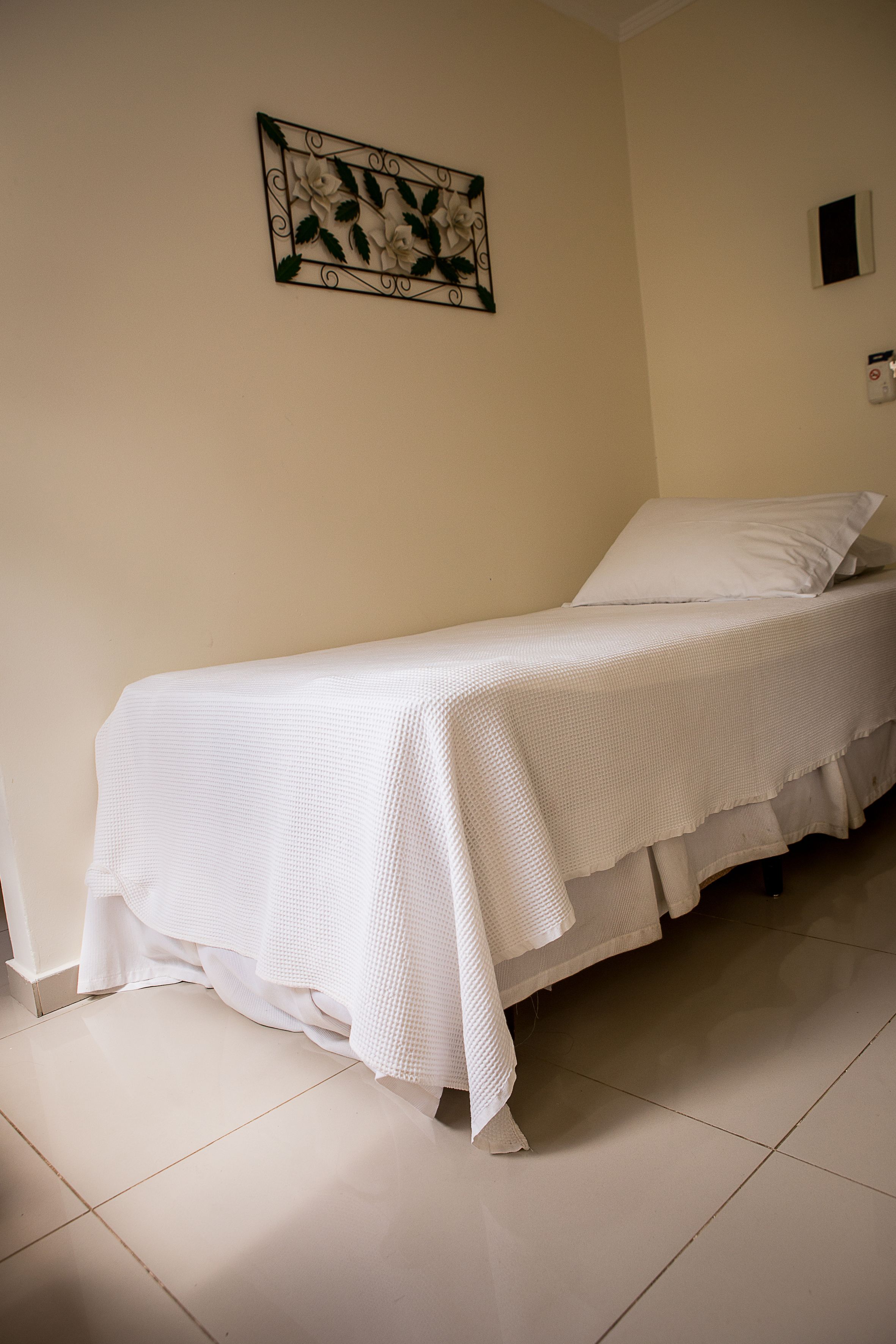 Uma cama de solteiro com um edredom branco em um quarto com parede lisa e uma obra de arte emoldurada acima.