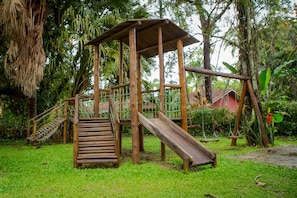 Una estructura de juego de madera con un tobogán y escaleras, rodeada de abundante vegetación.