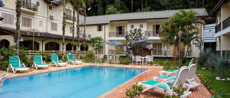 A pool area with lounge chairs and umbrellas, surrounded by a building with balconies and a garden.