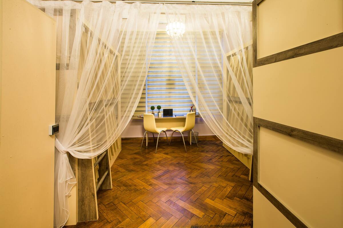 A room with a wooden herringbone floor, a small table with chairs, and sheer white curtains.