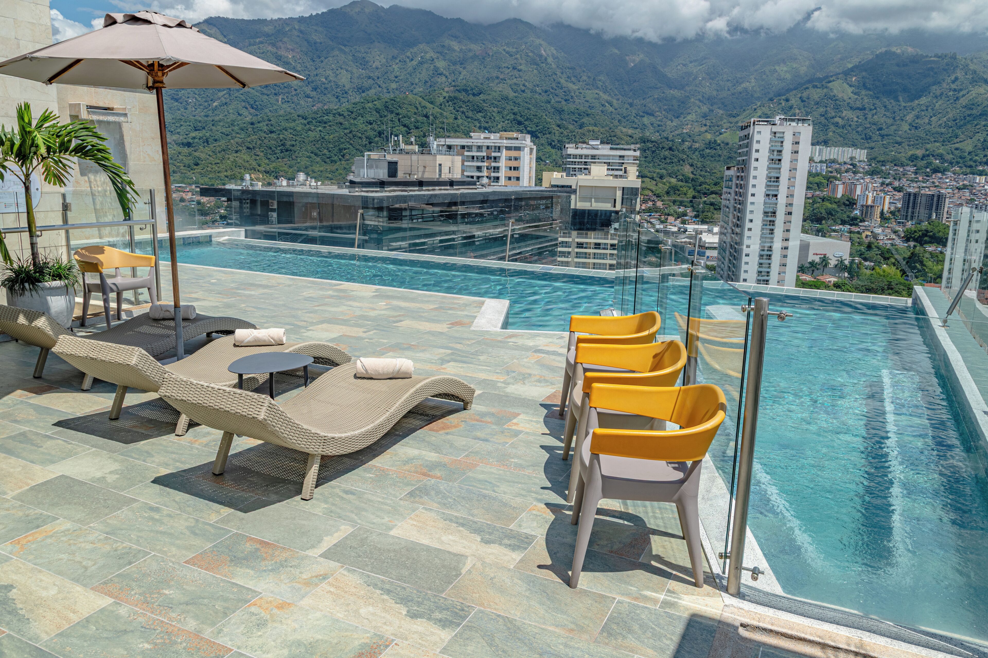 A rooftop infinity pool with lounge chairs and a table, overlooking a city and mountains.