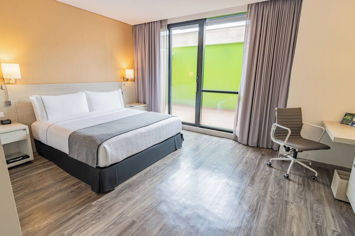 Een hotelkamer met een bed, nachtkastje, bureau, stoel en een grote glazen schuifpui naar een balkon.