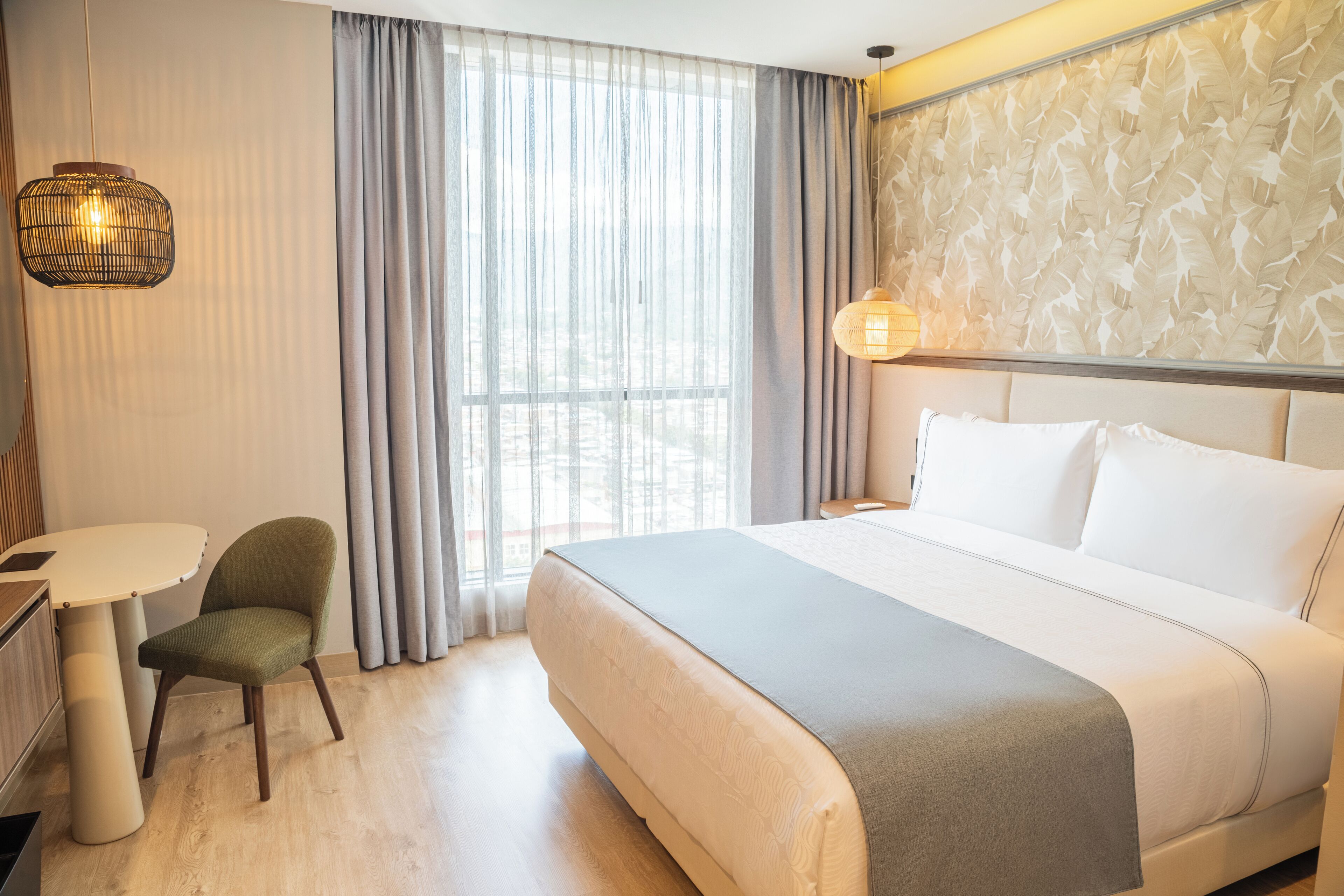 A hotel room with a large bed, a small table, a chair, and a window with curtains.