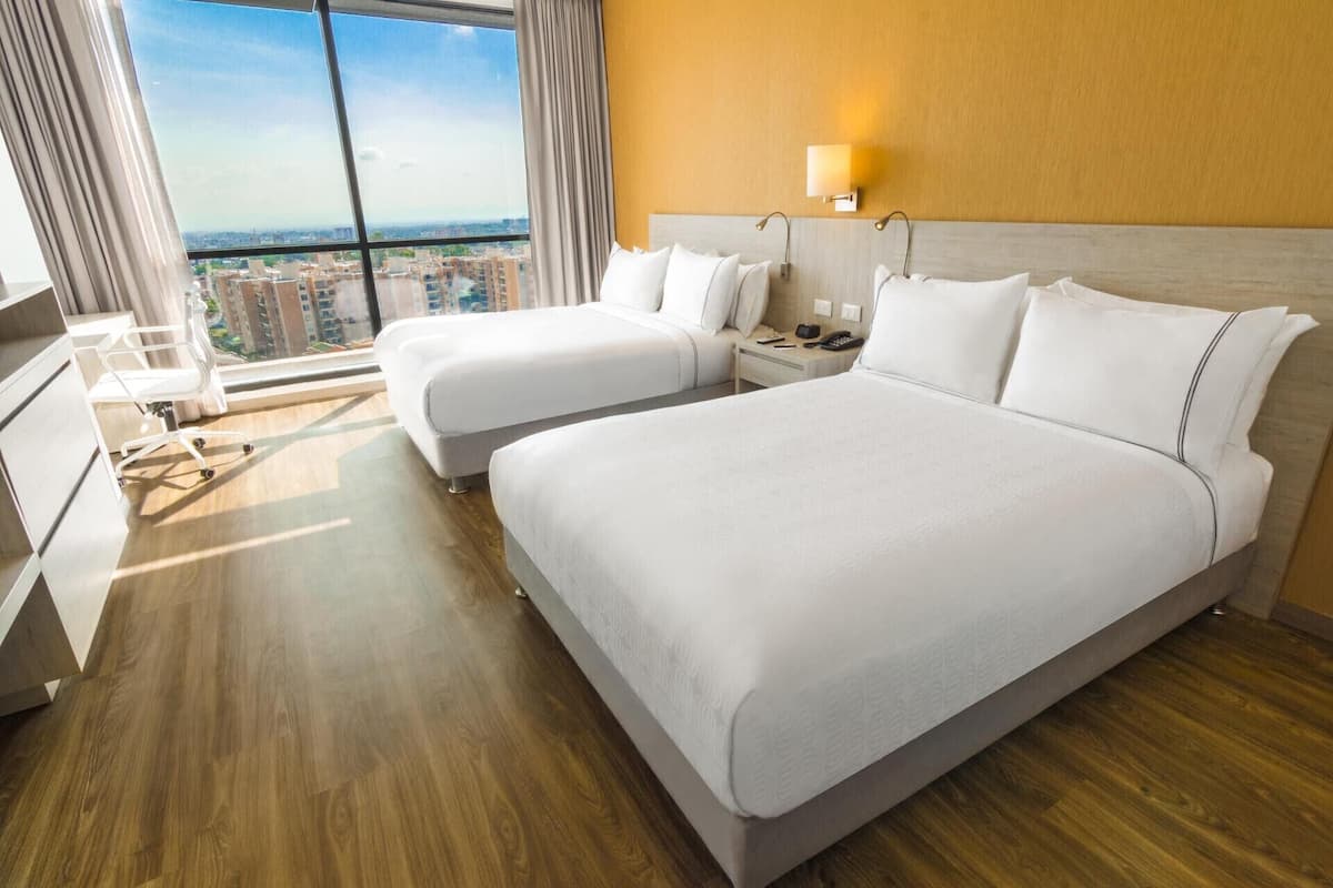 Hotelkamer met twee bedden, een bureau, een stoel, een nachtkastje en een groot raam met uitzicht op de stad.