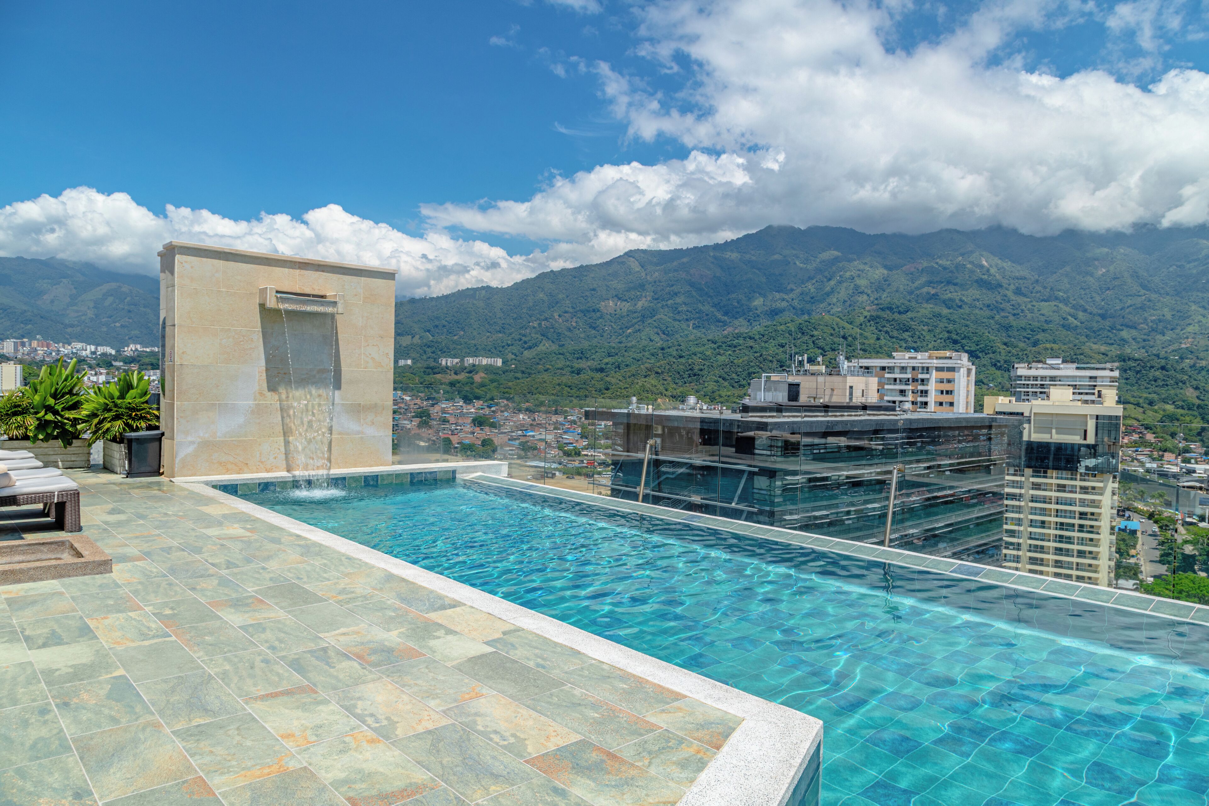 Una piscina infinita en la azotea con una cascada, rodeada de edificios modernos y un telón de fondo montañoso.