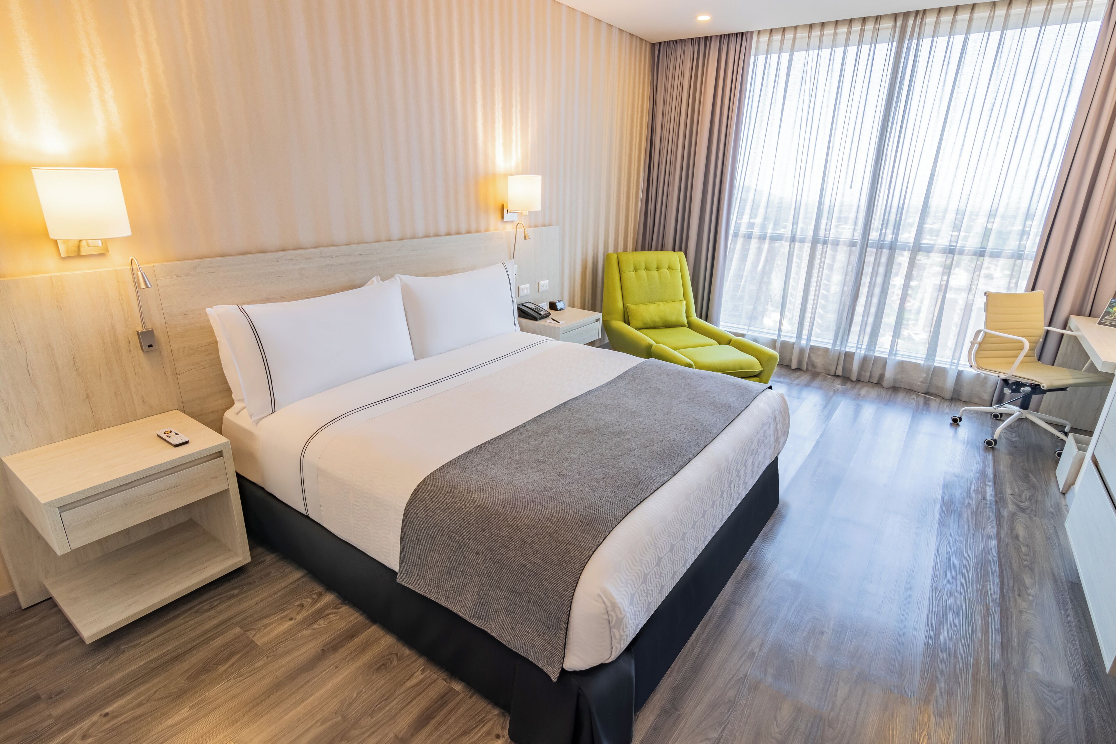 A hotel room with a bed, bedside tables, a desk, a chair, and a large window with curtains.