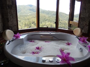 A hot tub with flower petals, wooden accents, and a view of a mountainous landscape through large windows. - Pousada do Sonho (Itatiaia)