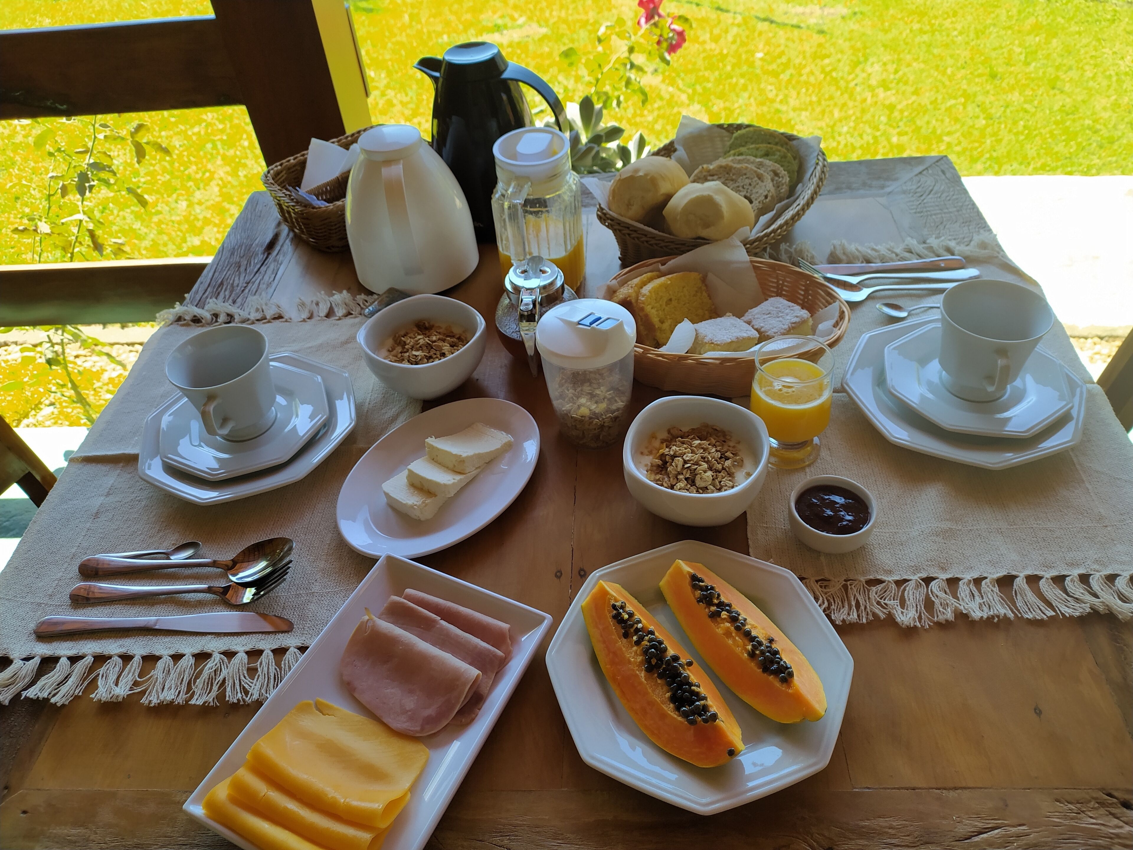 Una mesa de desayuno con diversos alimentos y bebidas, como pan, huevos y fruta, sobre una mesa de madera al aire libre.