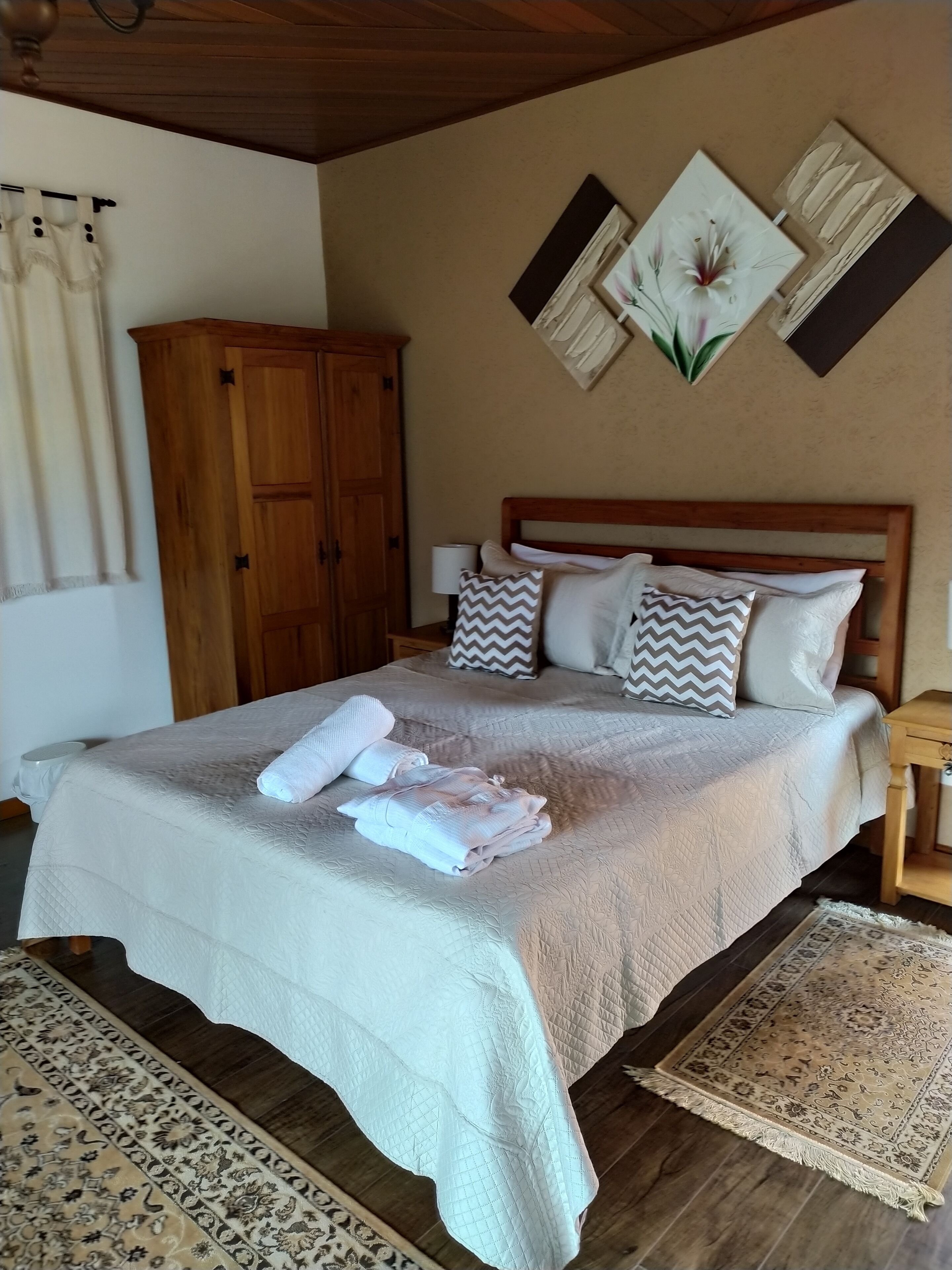 Un lit bien fait, recouvert d’une couette blanche, avec des oreillers et des serviettes pliées. Une armoire en bois se trouve à côté du lit, et un tapis à motifs est posé sur le sol.