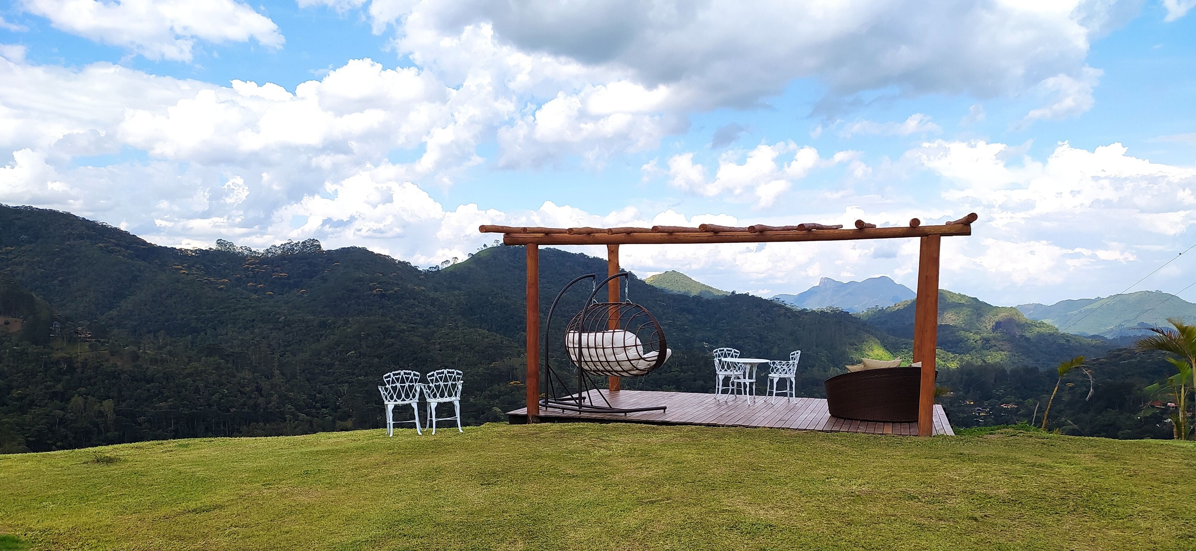 Una pérgola de madera con una silla colgante y dos sillas blancas, ubicada en una colina cubierta de césped con montañas al fondo.