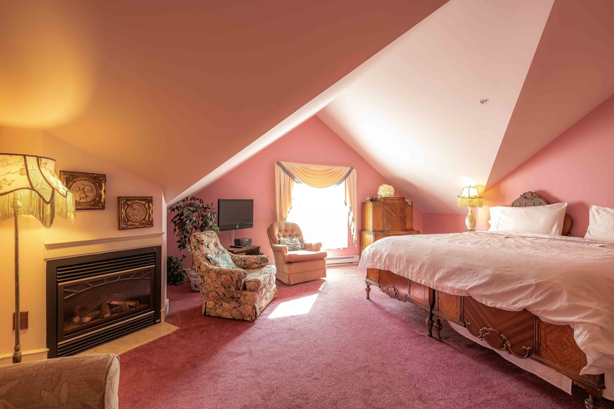 A bedroom with a sloped ceiling, a fireplace, a bed, a chair, and a television.