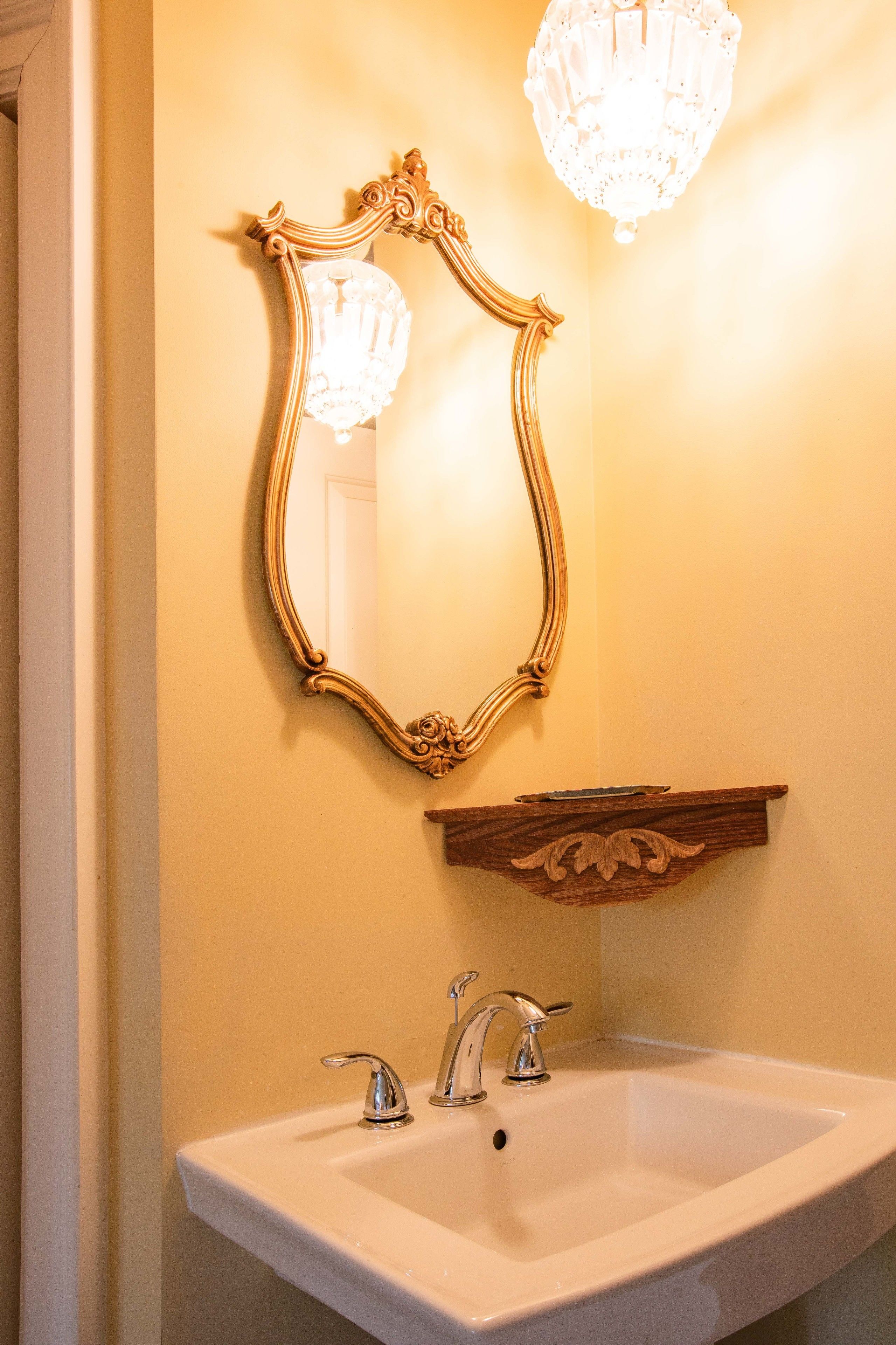 A bathroom with a white sink, two faucets, a gold-framed mirror, and a wooden shelf above the sink.