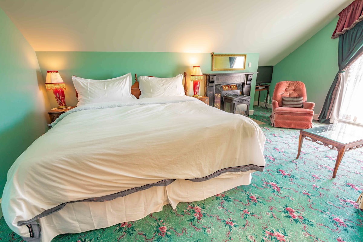 A large bed with white linens, a wooden chair, a small table, and a fireplace in a room with green walls and a patterned carpet.