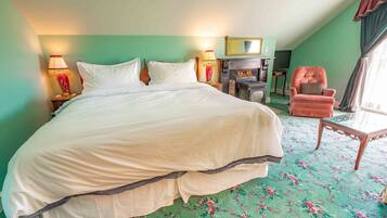 A large bed with white linens, a wooden chair, a small table, and a fireplace in a room with green walls and a patterned carpet.