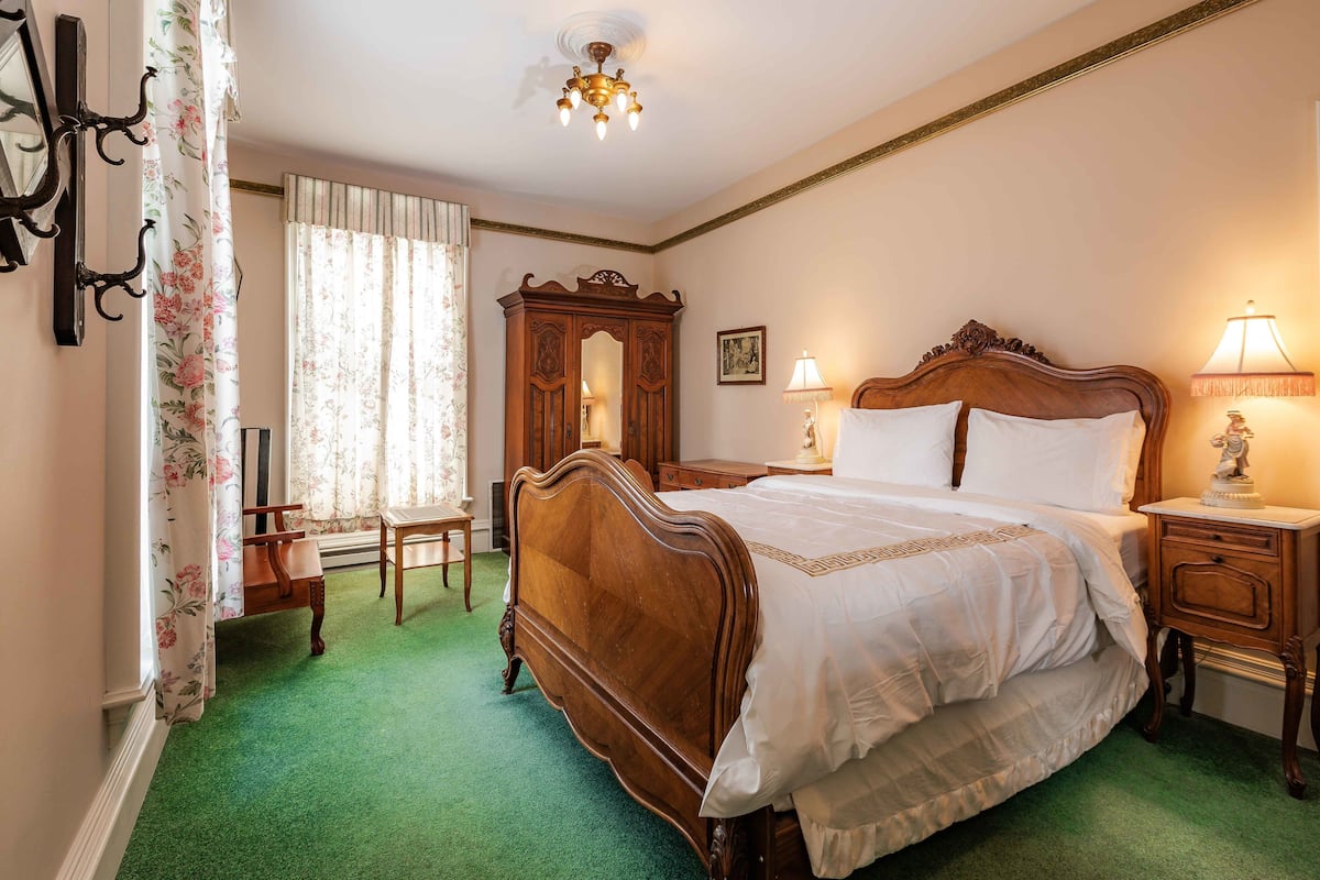 A bedroom with a wooden bed, two bedside tables with lamps, a wooden wardrobe, a chair, and a window with curtains.