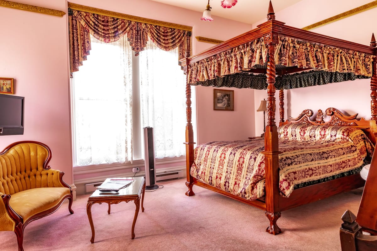 A four-poster bed with a patterned bedspread, a yellow armchair, a small table, and a large window with curtains.