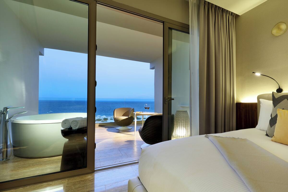 A hotel room with a large bed, a glass door leading to a balcony with a view of the sea, a bathtub, and a table with a glass of wine.
