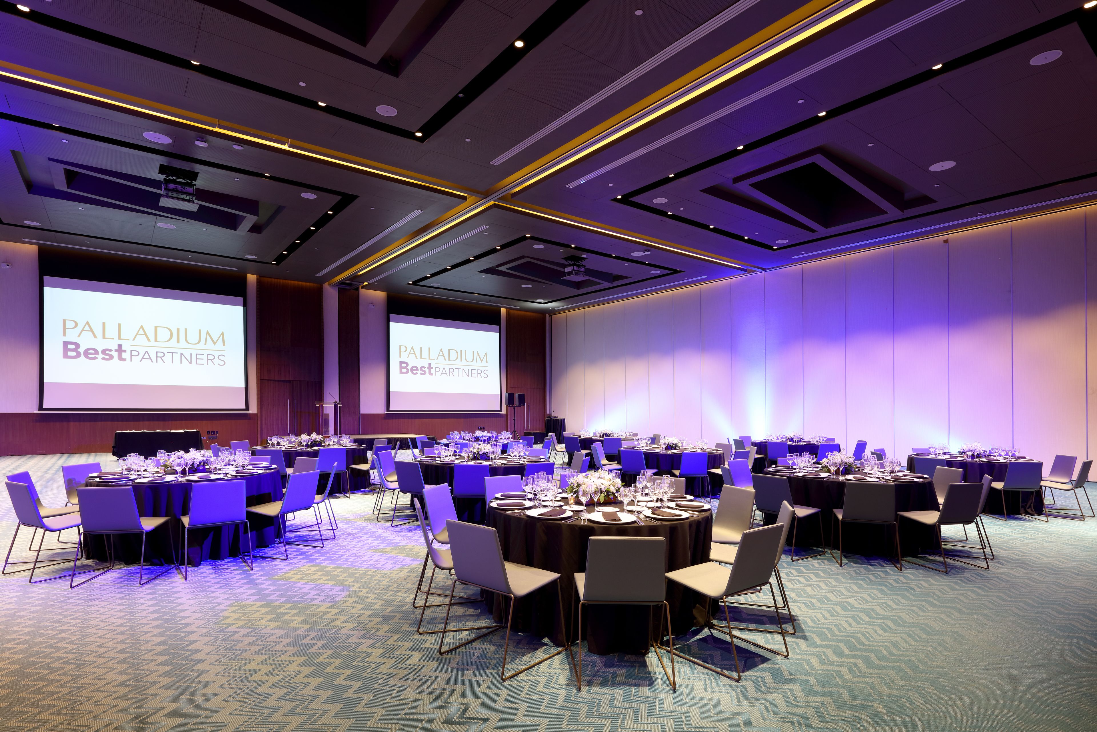 Un salón de conferencias con varias mesas redondas, sillas y una pantalla grande que muestra 'Palladium Best Partners'.