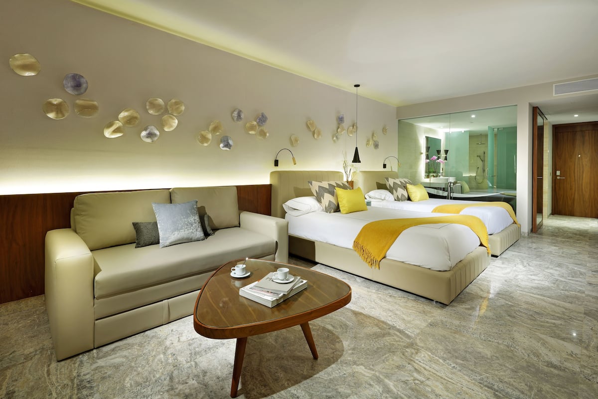 A modern hotel room with a sofa, a bed with a yellow throw, a coffee table with a book, and a decorative wall with stone-like elements.