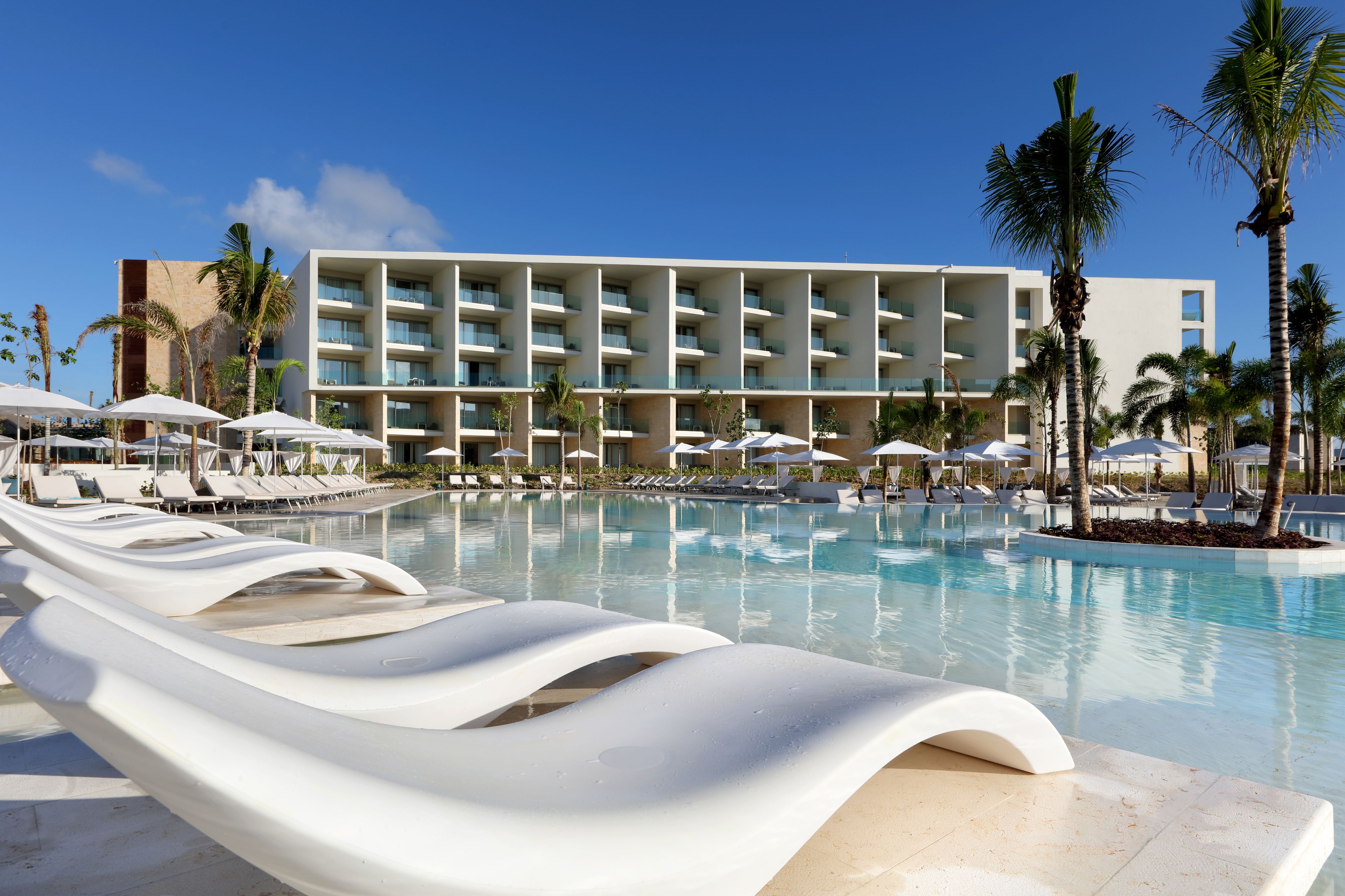 A hotel pool area with white lounge chairs and umbrellas, surrounded by palm trees and a modern building with multiple balconies.