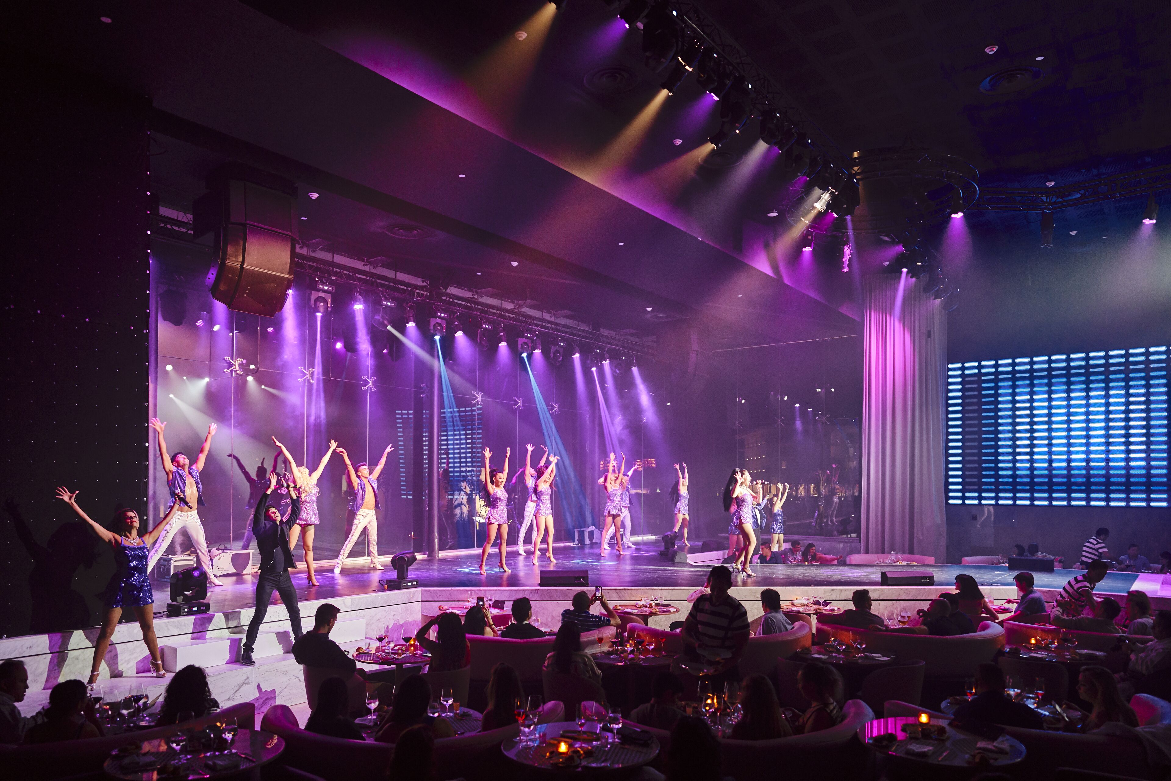 A stage performance with dancers in a dimly lit venue, audience seated at tables, and a large screen in the background.