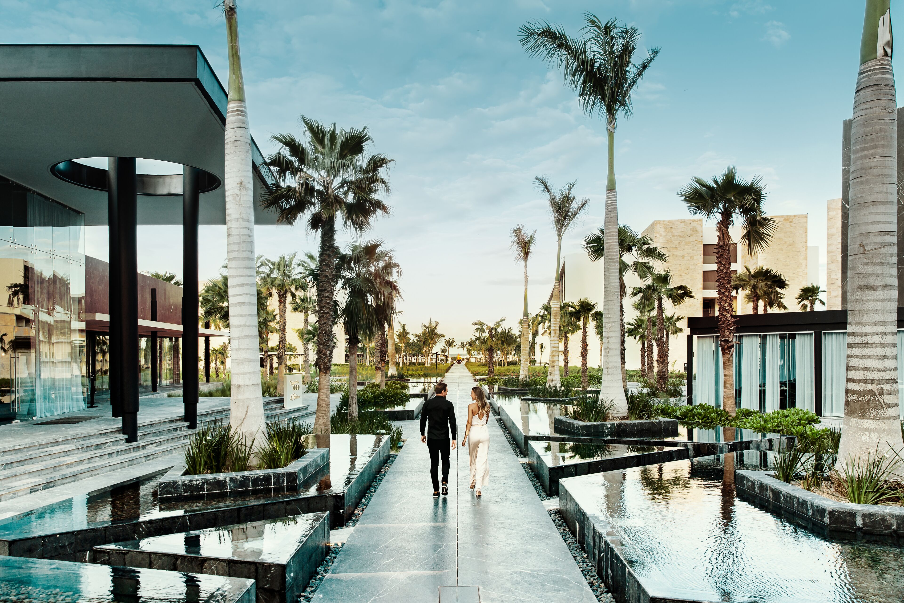 A couple walking on a water feature pathway surrounded by palm trees and modern buildings.