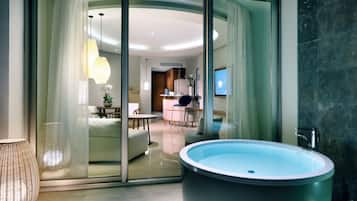 Un baño moderno con una bañera grande, cabina de ducha con mampara de vidrio y una zona de comedor visible a través de las puertas de cristal.