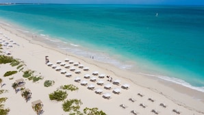 A beach with rows of sun loungers and umbrellas, a sandy shore, and a clear blue sea.