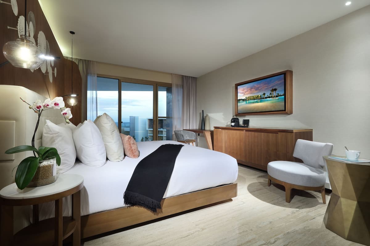 A modern hotel room with a large bed, a desk, a chair, a small table with a plant, and a large window with a city view.