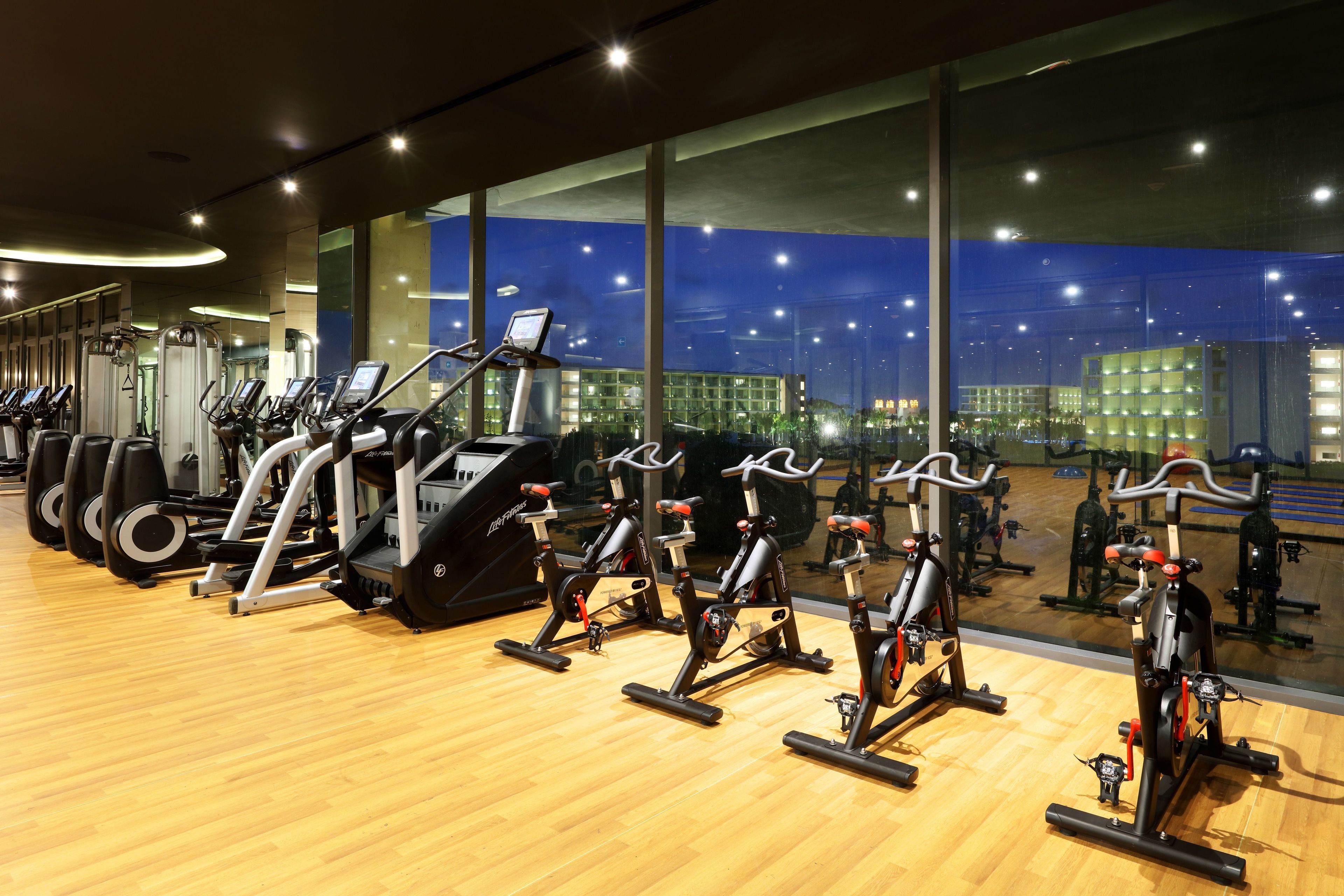 Een sportschool met diverse fitnessapparaten, grote ramen en een uitzicht over de stad 's nachts.