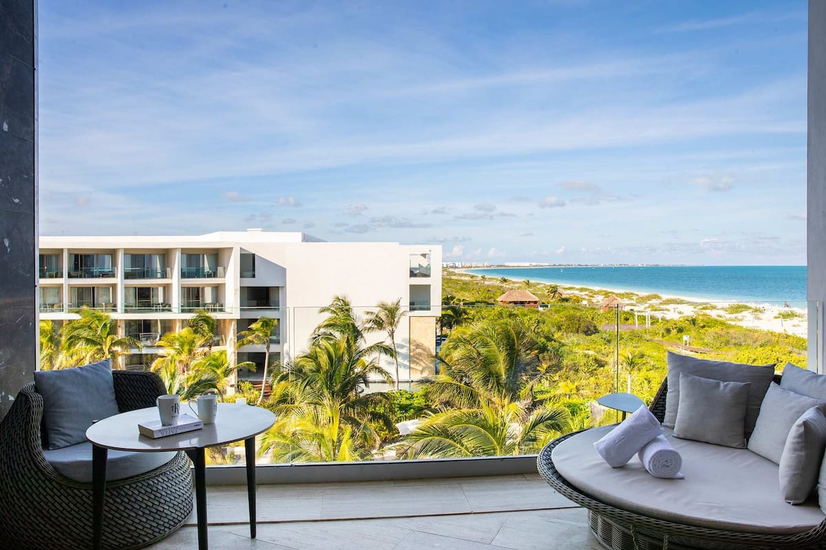 A balcony with a view of a beach and a modern building. There are wicker chairs and a small table with a book and a mug.