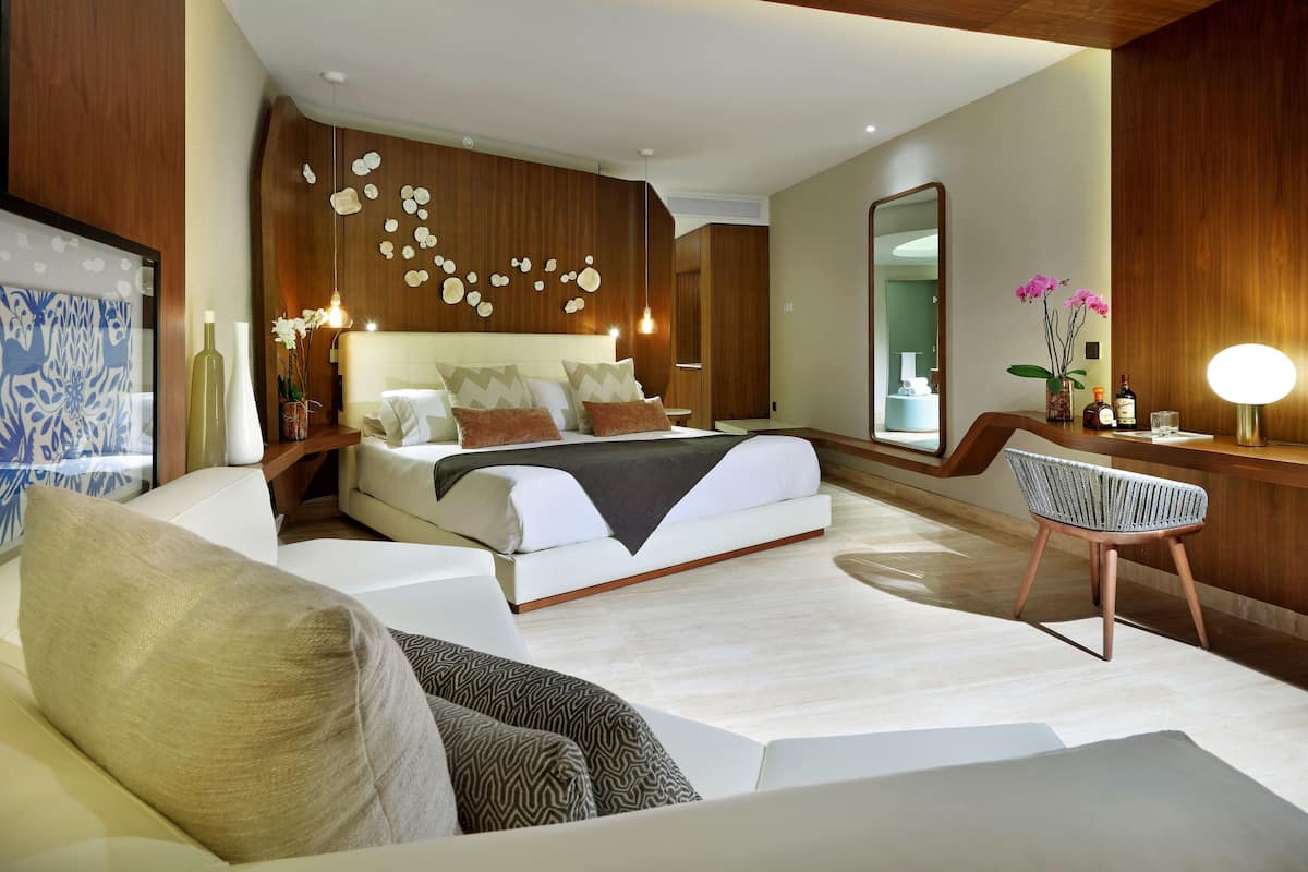 A modern bedroom with a large bed, a sitting area with a chair, and a vanity with a mirror.